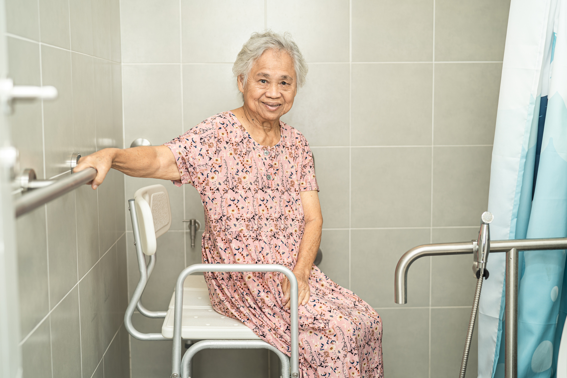 Na obrazku widoczna jest uśmiechnięta seniorka w łazience przystosowanej dla seniorów, pełnej funkcjonalnych udogodnień i elementów zapewniających bezpieczeństwo. Poręcze, podpórki i antypoślizgowe powierzchnie pozwalają jej cieszyć się niezależnością i komfortem podczas codziennych czynności higienicznych. Ten obrazek ukazuje znaczenie odpowiedniego projektowania łazienki dla starszych osób, które mają możliwość korzystania z przestrzeni w sposób wygodny i bezpieczny